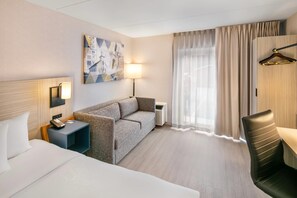 Une chambre d’hôtel comprenant un lit, un canapé, un bureau, une chaise, une lampe, un radiateur, une fenêtre avec des rideaux et une œuvre d’art murale.