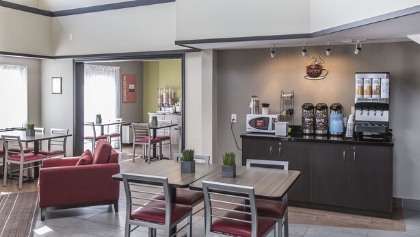 A modern dining area with red upholstered chairs and a wooden table, a kitchen area with a coffee machine and dispensers, and a view of the adjacent room.
