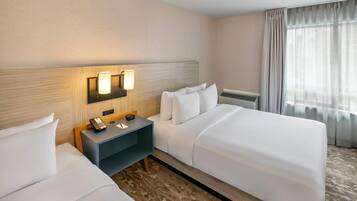 A hotel room with two beds, a nightstand with a phone and a clock, a wall-mounted lamp, and a window with curtains.