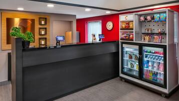 Uma área de recepção com balcão preto, um relógio, uma tela digital, um refrigerador abastecido com bebidas e uma planta.