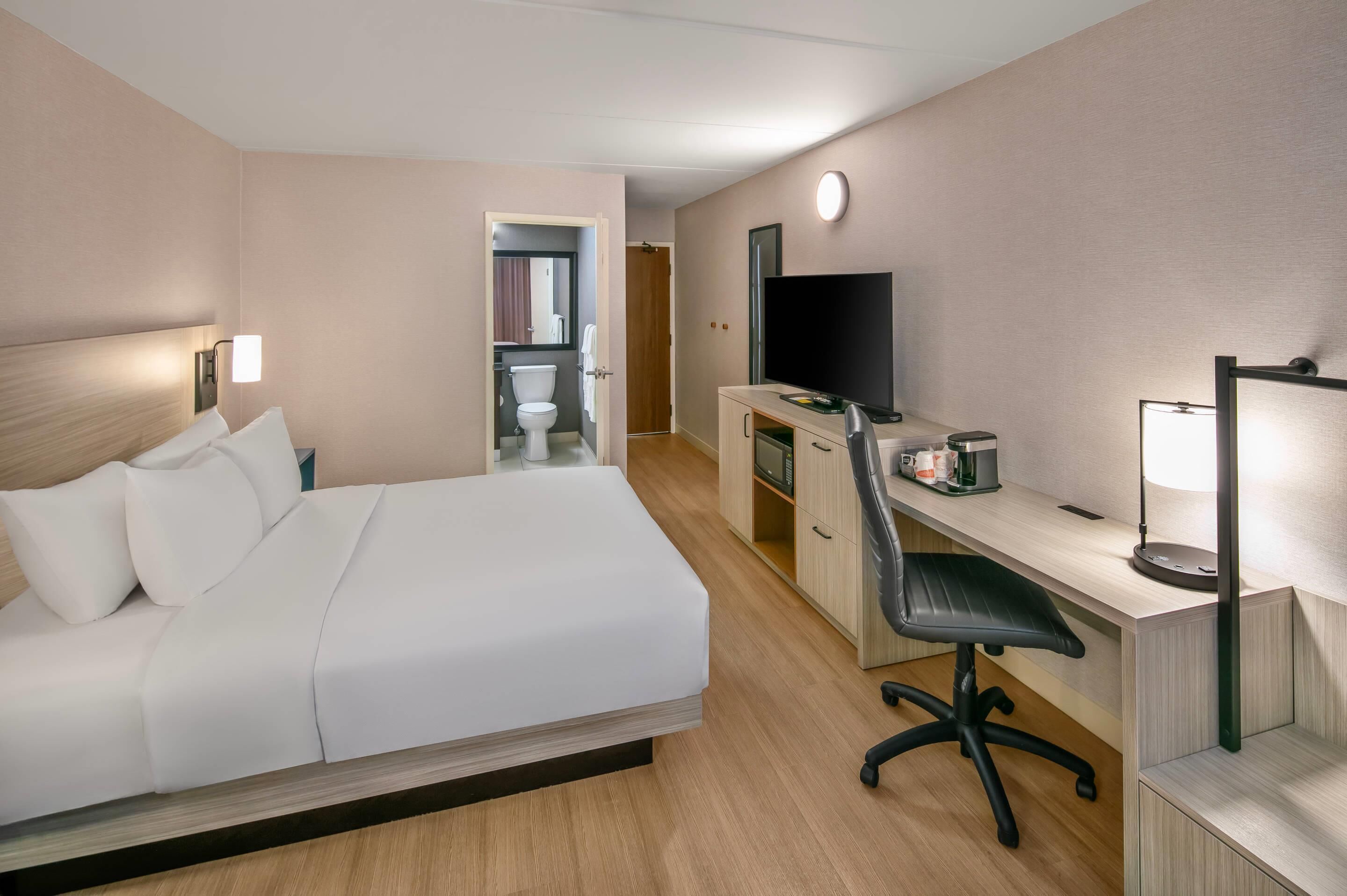 Ein Hotelzimmer mit Bett, Schreibtisch, Stuhl, Fernseher und einem durchgegangene Tür erkennbaren Badezimmer.