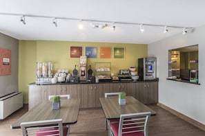 A modern kitchen with a central island, equipped with coffee machines, a microwave, and a refrigerator. There are two dining tables with chairs, and the walls are decorated with colorful posters.