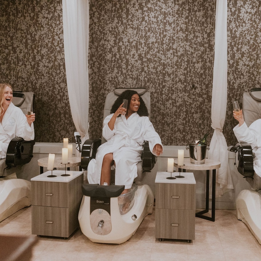 Three individuals in a spa setting, each seated in a foot massage chair with a foot bath, surrounded by candles and small tables.