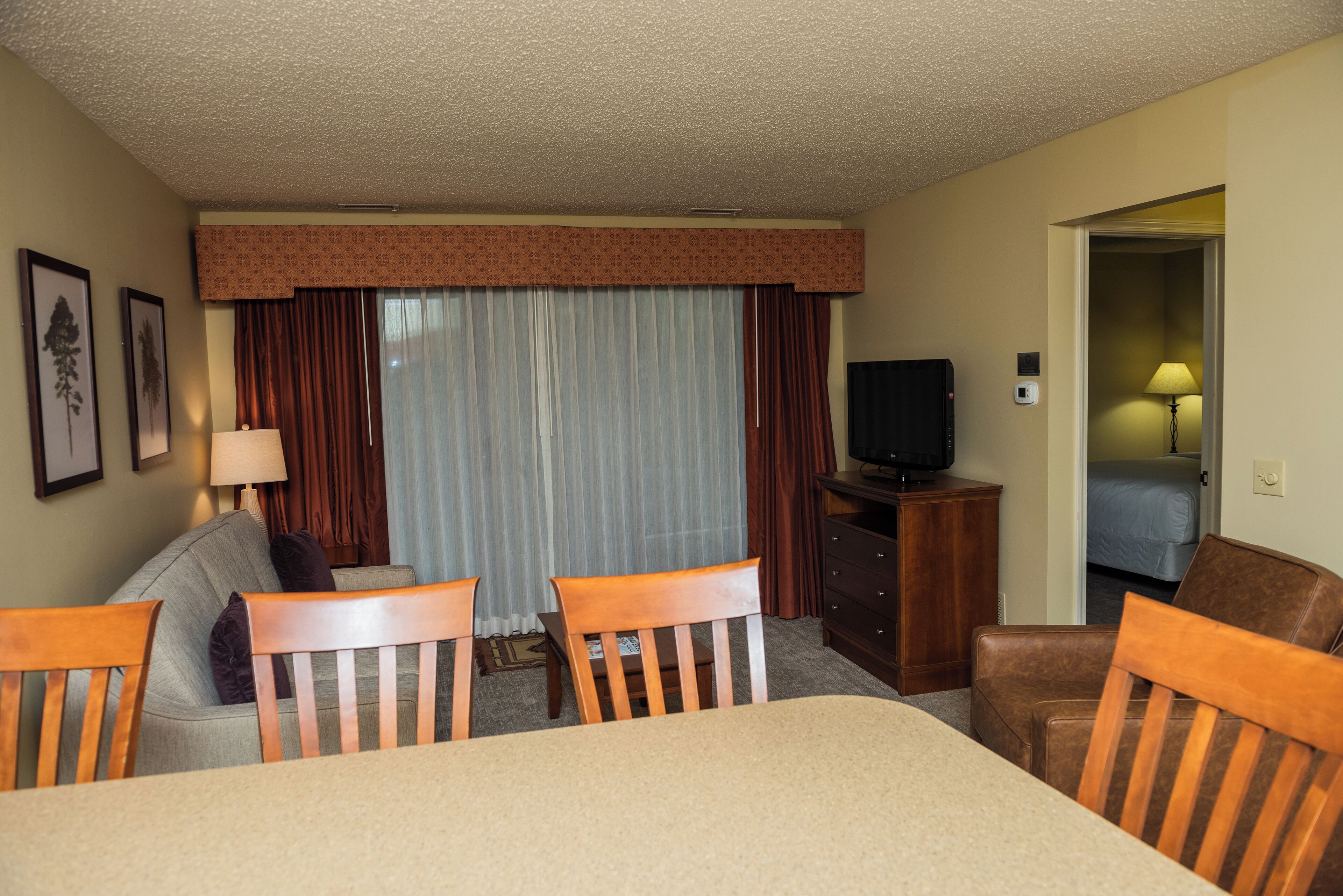 A hotel room with a dining table, two wooden chairs, a sofa, a television, a lamp, and a window with curtains.