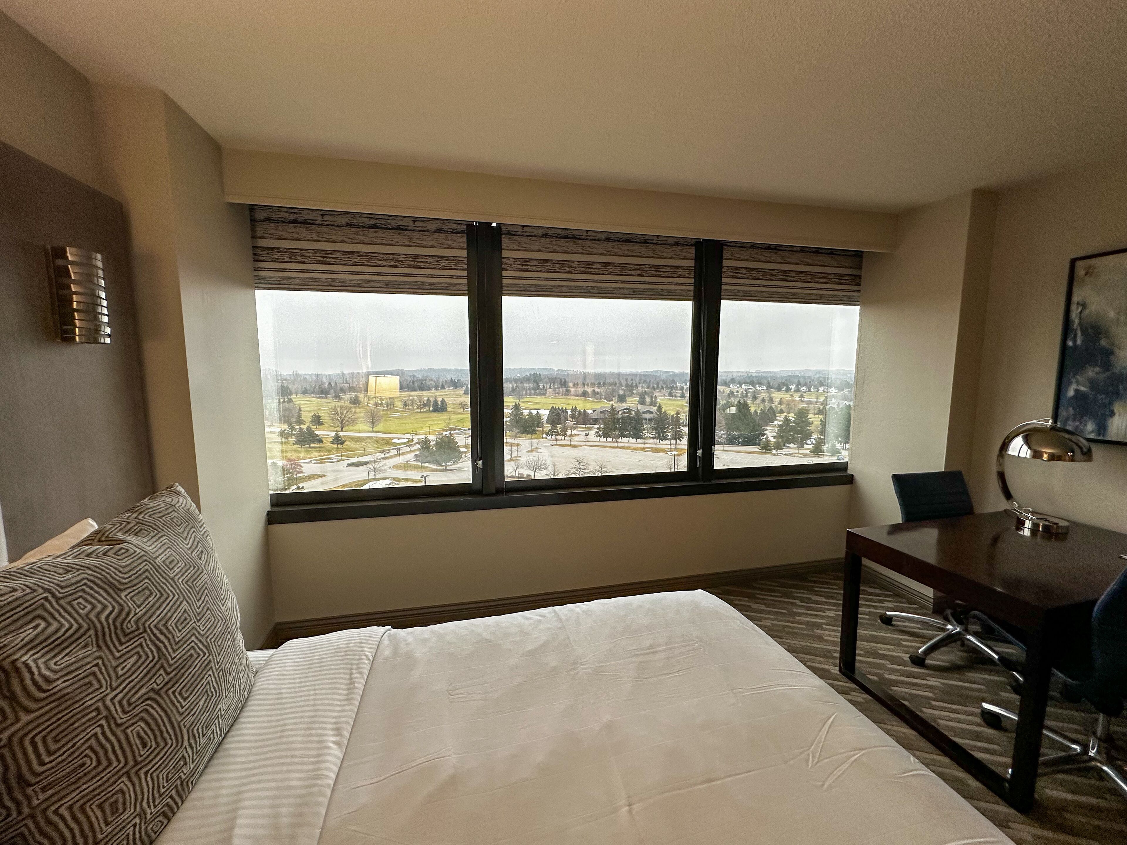 A hotel room with a large window, a bed, a desk, and a chair.