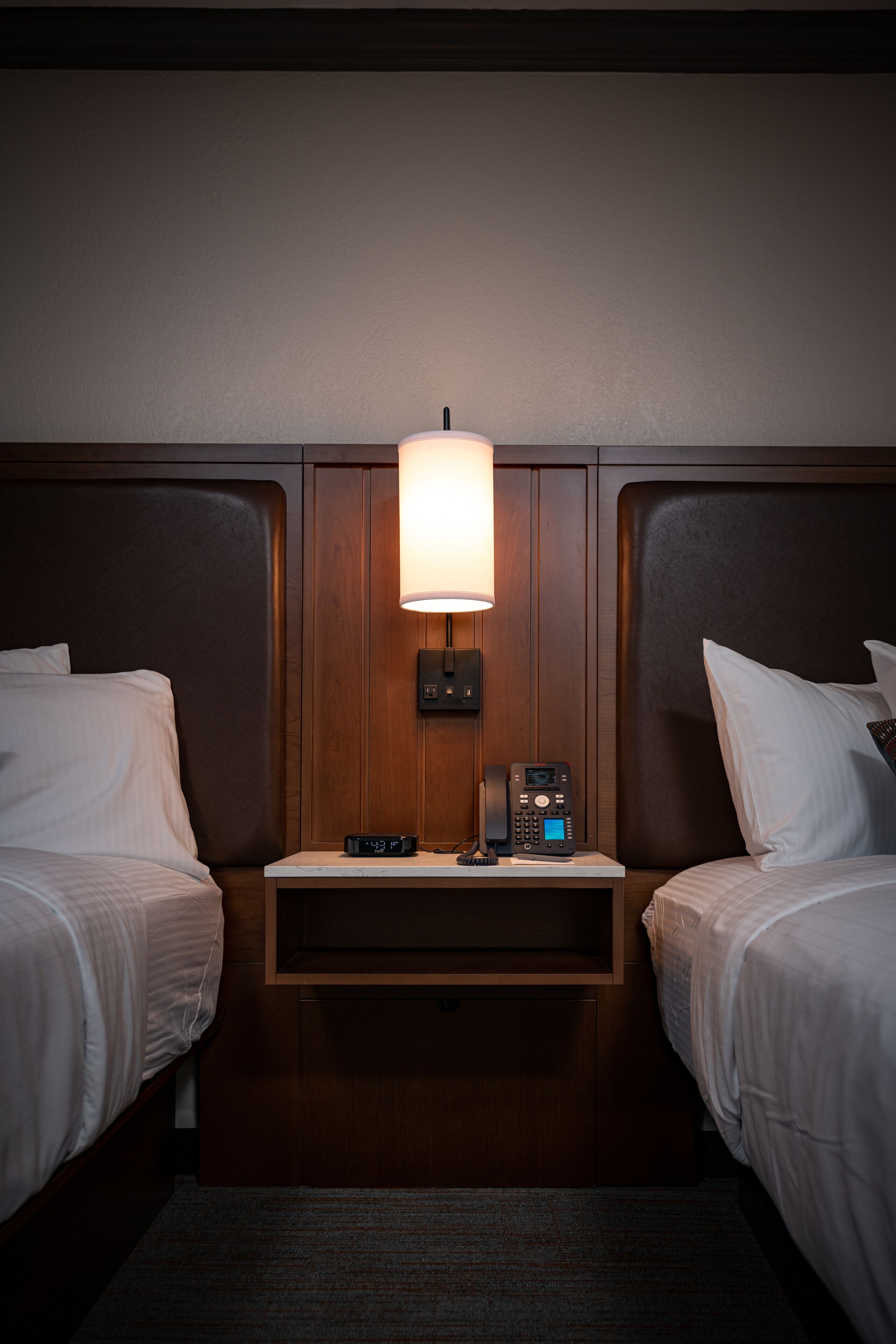 A hotel room with two beds, a bedside table with a lamp, and a telephone.