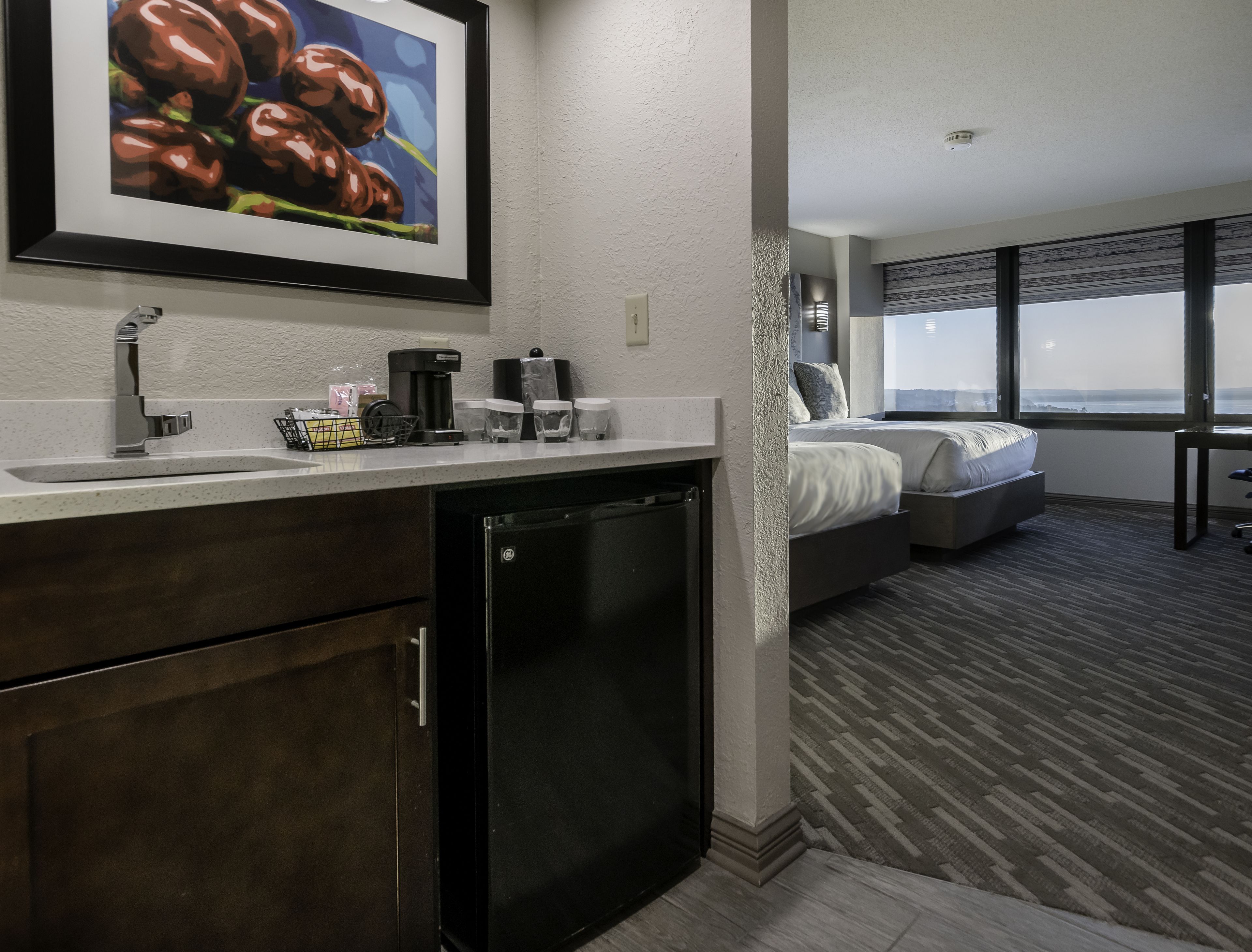 A hotel room with a kitchenette, a bed, a large window, and a view of the city.