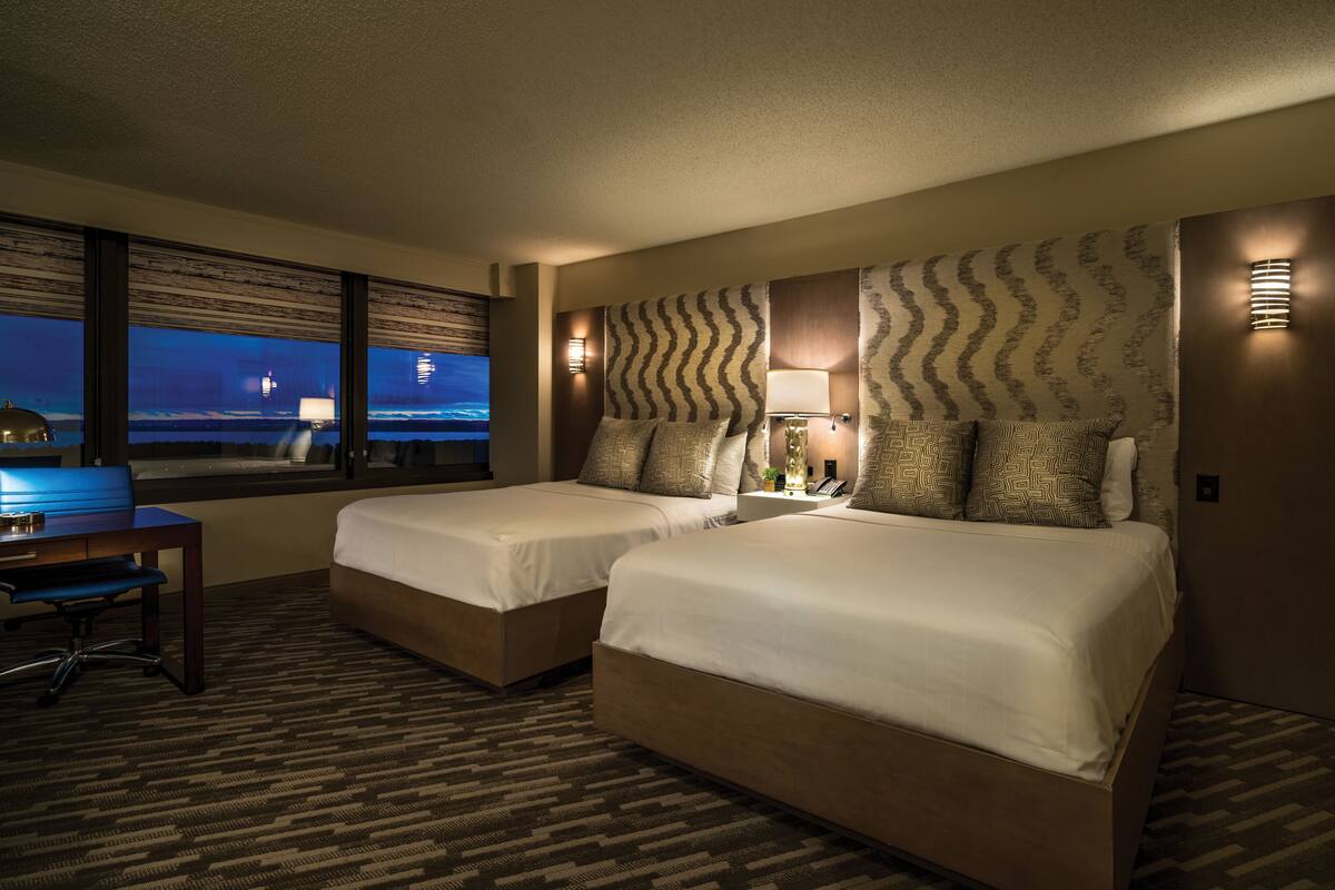 A hotel room with two beds, a desk, and a view of the cityscape at night.