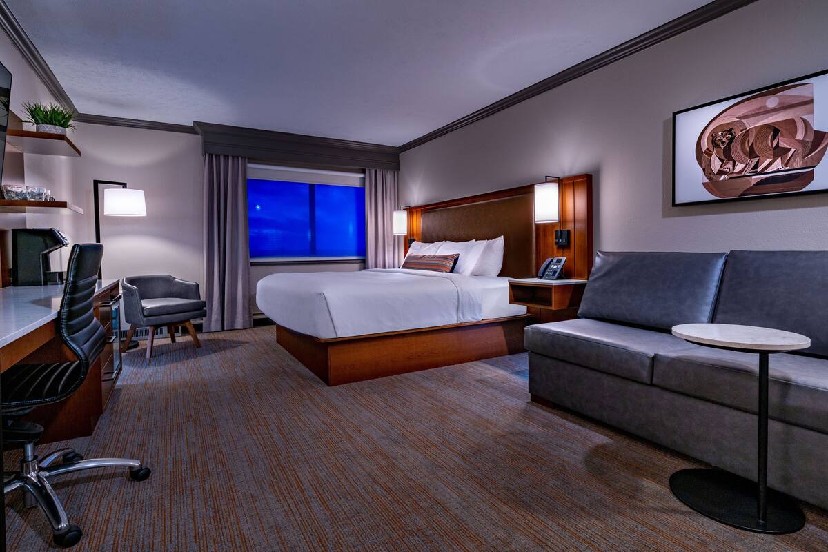 A hotel room with a bed, a sofa, a desk with a chair, a small table, and a large window with a view of the night sky.