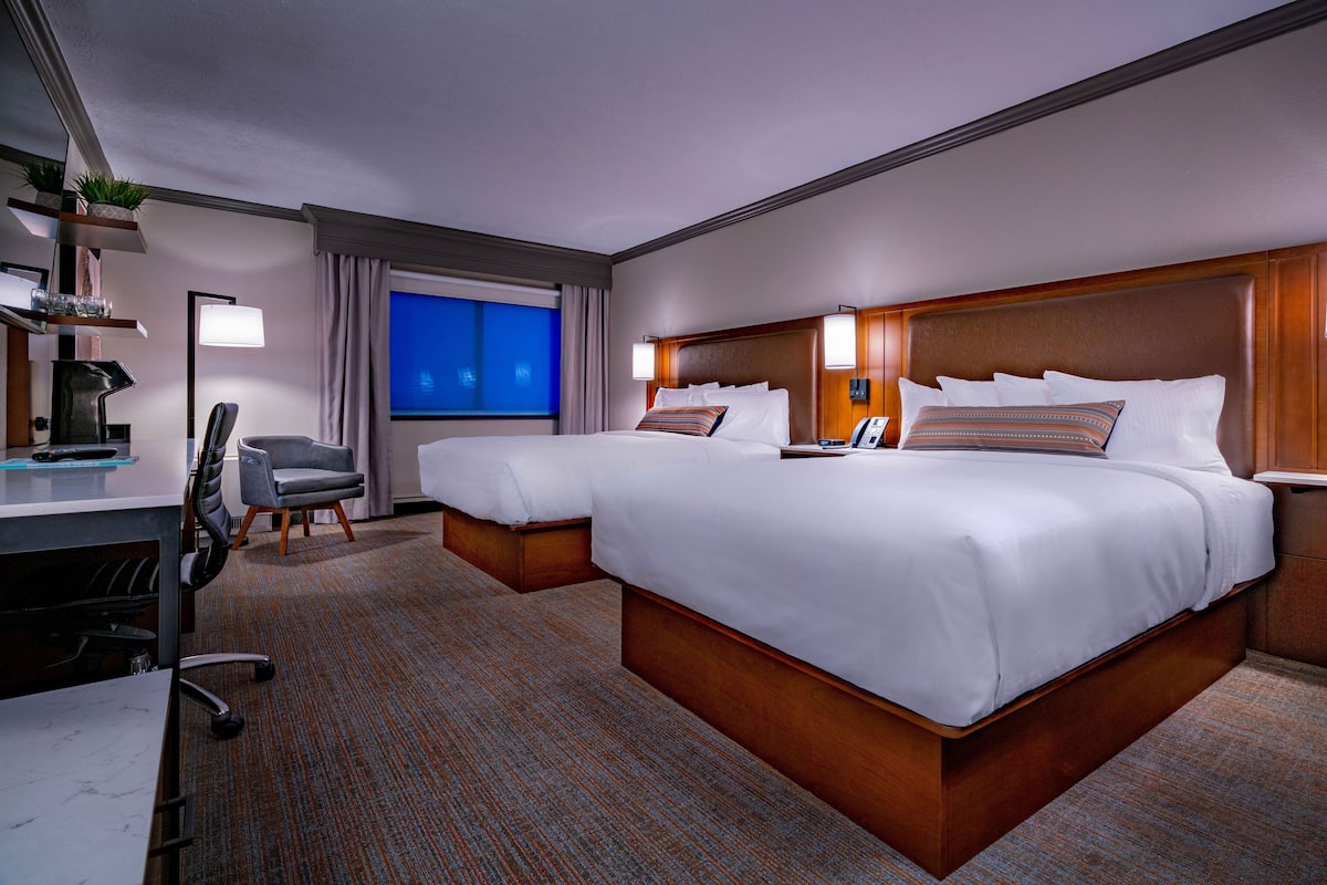 A hotel room with two beds, a desk, a chair, a lamp, and a window with a view.