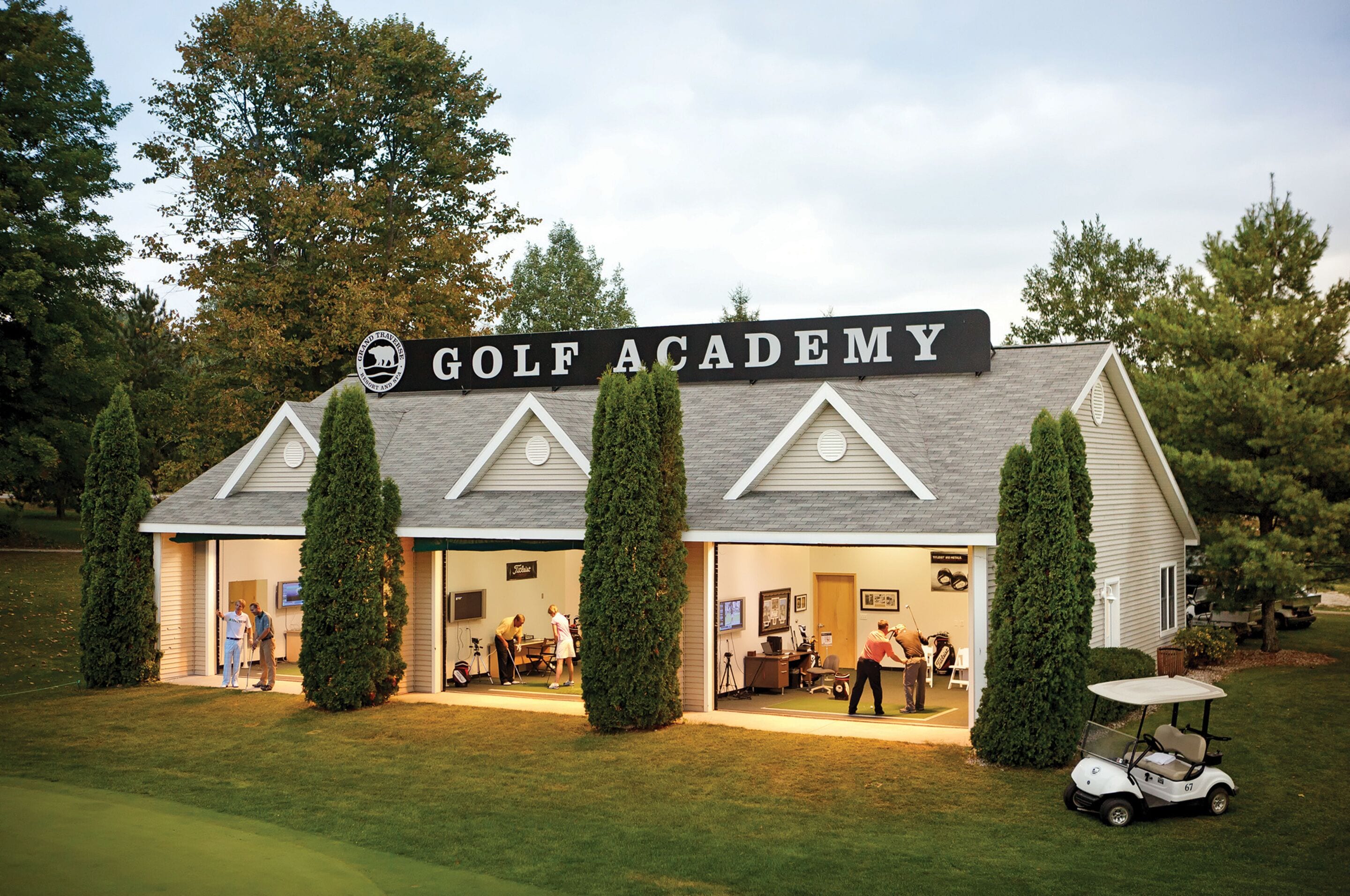 Un bâtiment abritant une académie de golf, avec une enseigne, plusieurs portes et un chariot de golf à l’extérieur.