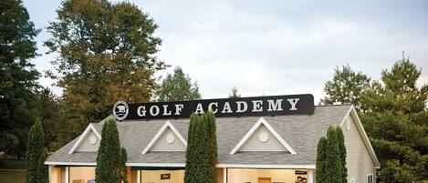 Un bâtiment abritant une académie de golf, avec une enseigne, plusieurs portes et un chariot de golf à l’extérieur.