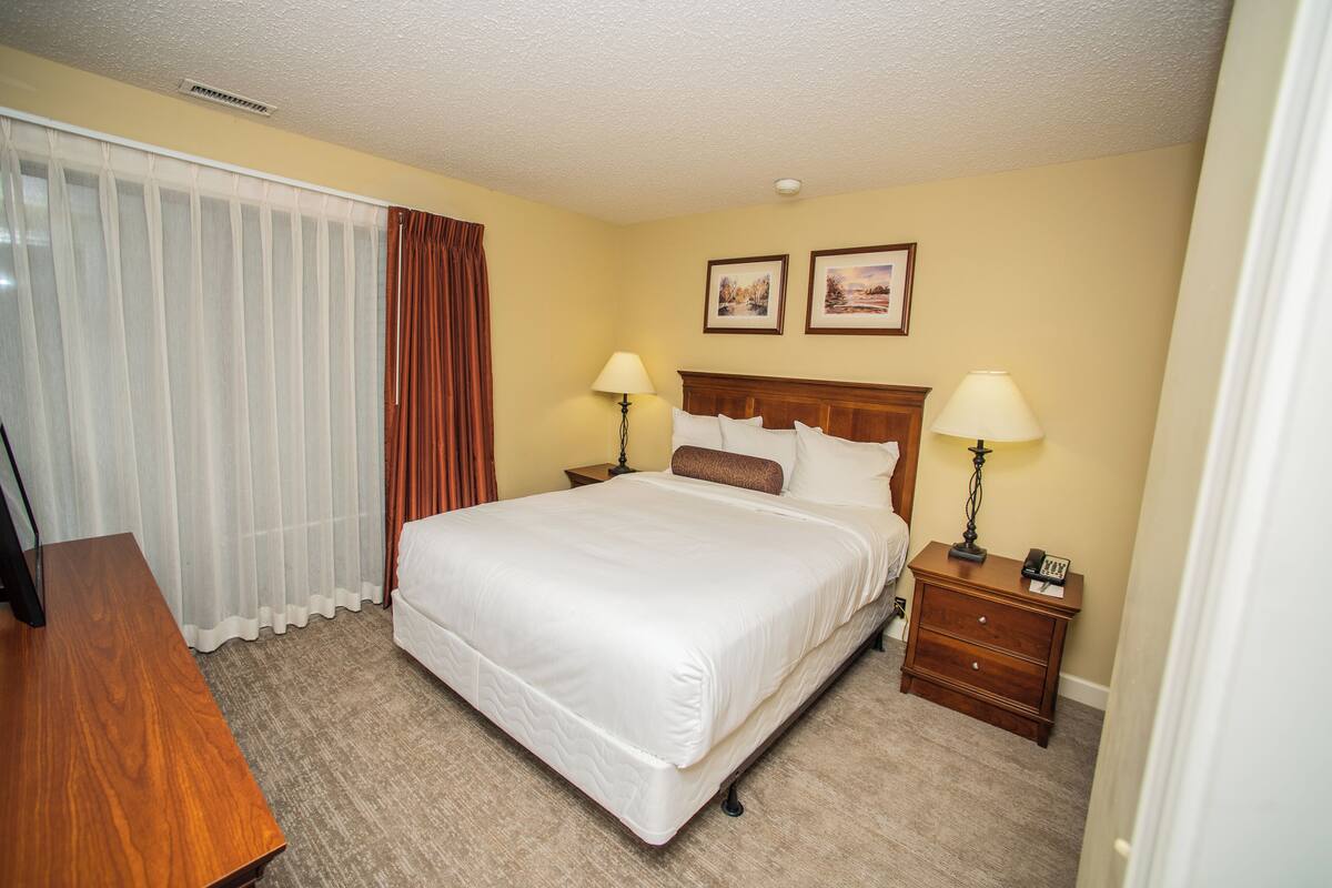 A hotel room with a large bed, two bedside tables with lamps, a wooden desk, and a window with curtains.