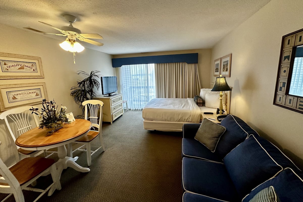 A compact hotel room with a bed, a blue sofa, a dining table with chairs, a TV, and a ceiling fan.