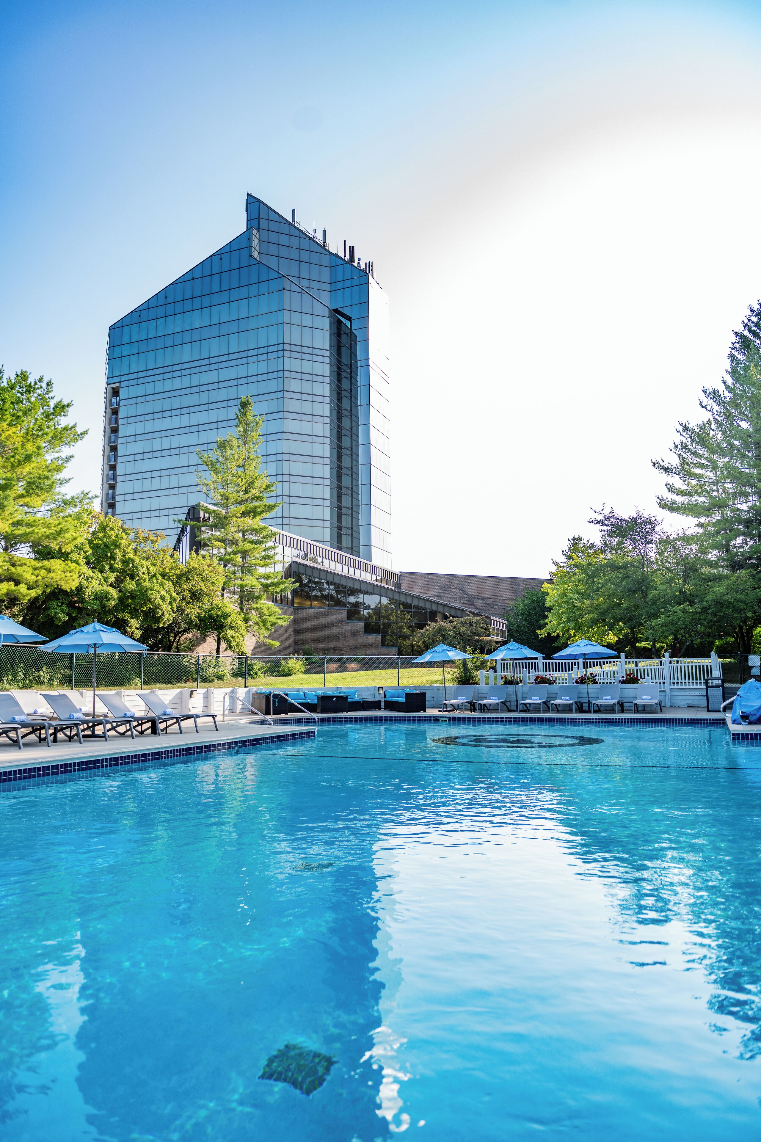 A swimming pool with lounge chairs and umbrellas, surrounded by trees and a tall building in the background. - Grand Traverse Resort And Spa (Williamsburg)