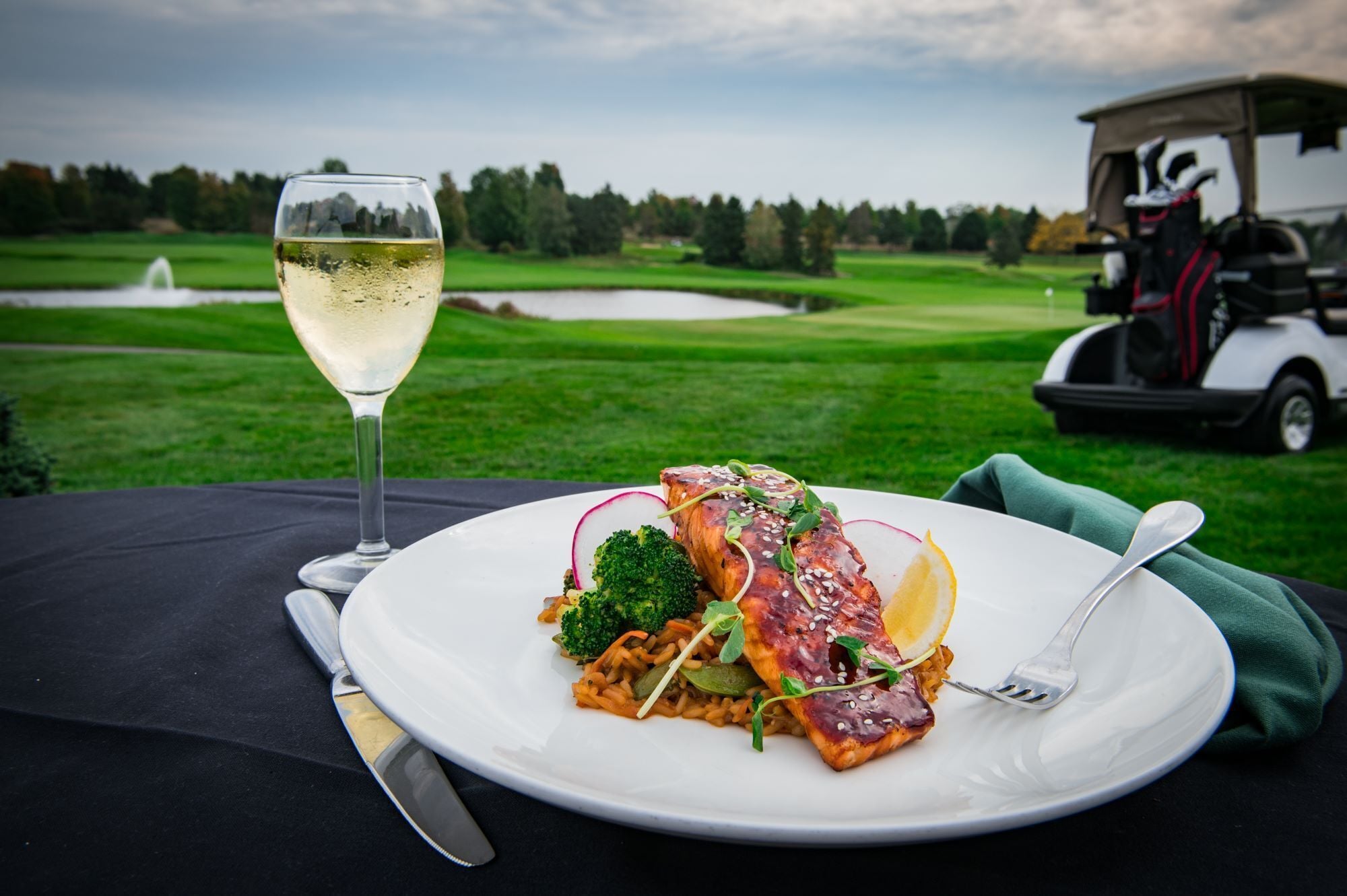 A meal with salmon, broccoli, and radish on a white plate, a glass of white wine, and a golf cart in the background.