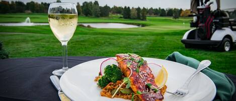 Un repas composé de saumon, de brocoli et de radis, servi dans une assiette blanche, accompagné d’un verre de vin blanc, et avec un chariot de golf en arrière-plan.