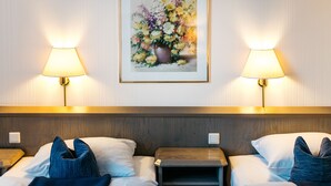 Habitación de hotel con dos camas, cada una con almohada y manta. Dos mesitas de noche, una al lado de cada cama. Un cuadro enmarcado con flores cuelga de la pared sobre las camas.