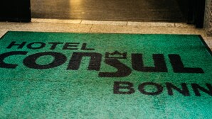 Zona de recepción de un hotel, con un felpudo verde que dice "HOTEL CONSUL BONN" y una puerta de vidrio que da al interior.