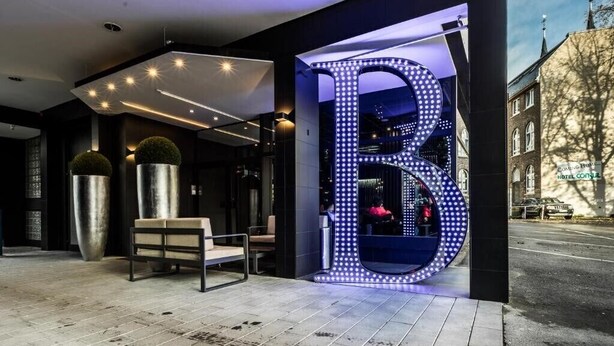 Una entrada de hotel moderna con un llamativo letrero iluminado con la letra 'B', una zona de estar con un banco y vista a la calle.