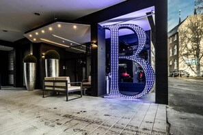 A modern hotel entrance with a prominent illuminated 'B' sign, a seating area with a bench, and a visible street view. - Consul (Bonn)