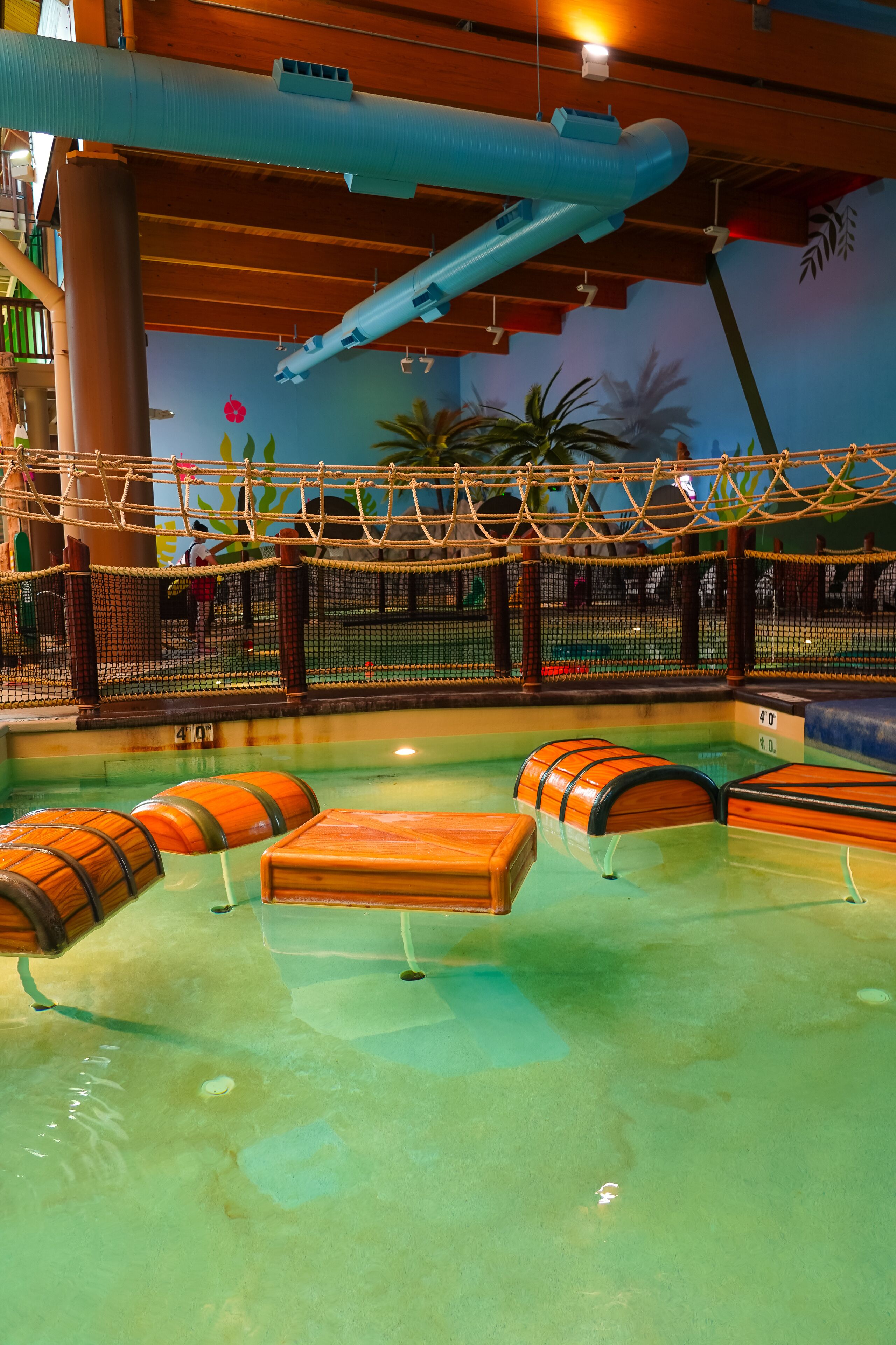 An indoor water play area with a rope bridge, wooden barrels, and a netted enclosure.