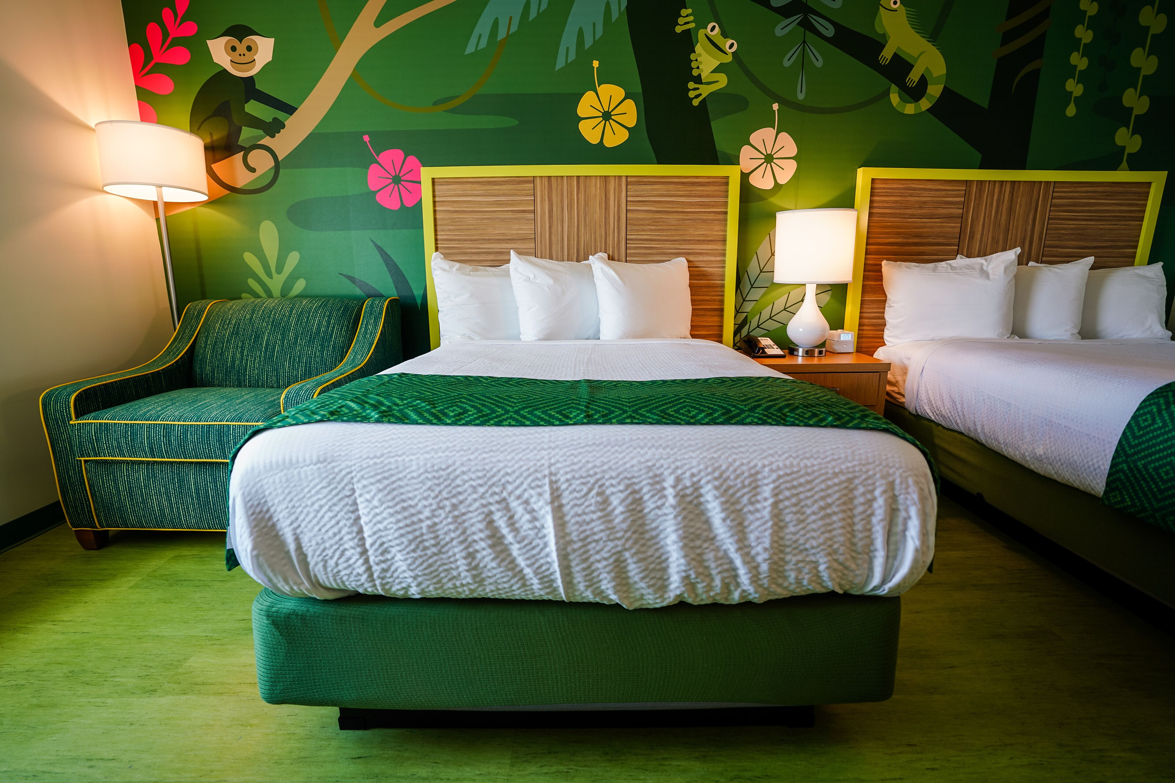 A hotel room with two beds, a green sofa, and a mural with animal and flower designs.