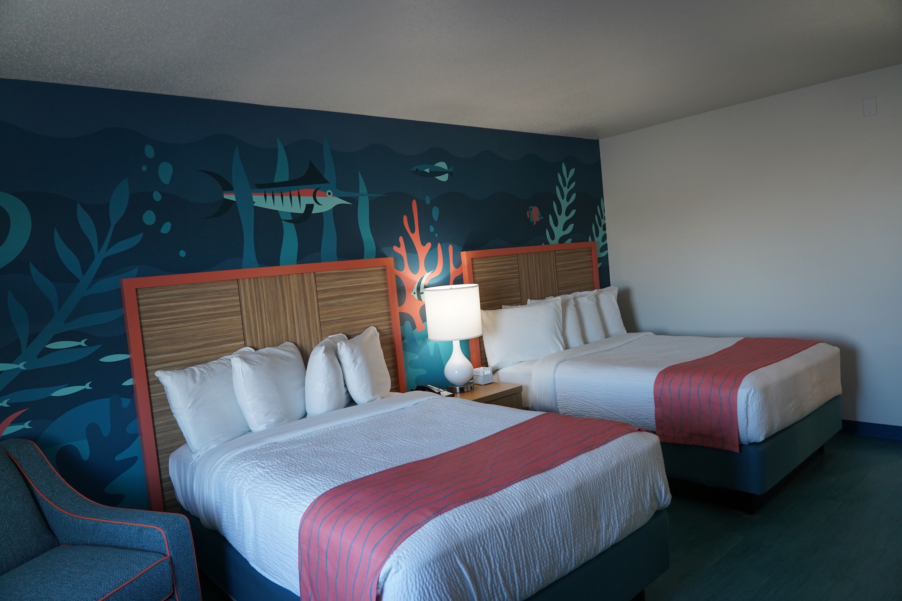 A hotel room with two beds, a nightstand with a lamp, and an underwater-themed mural on the wall.