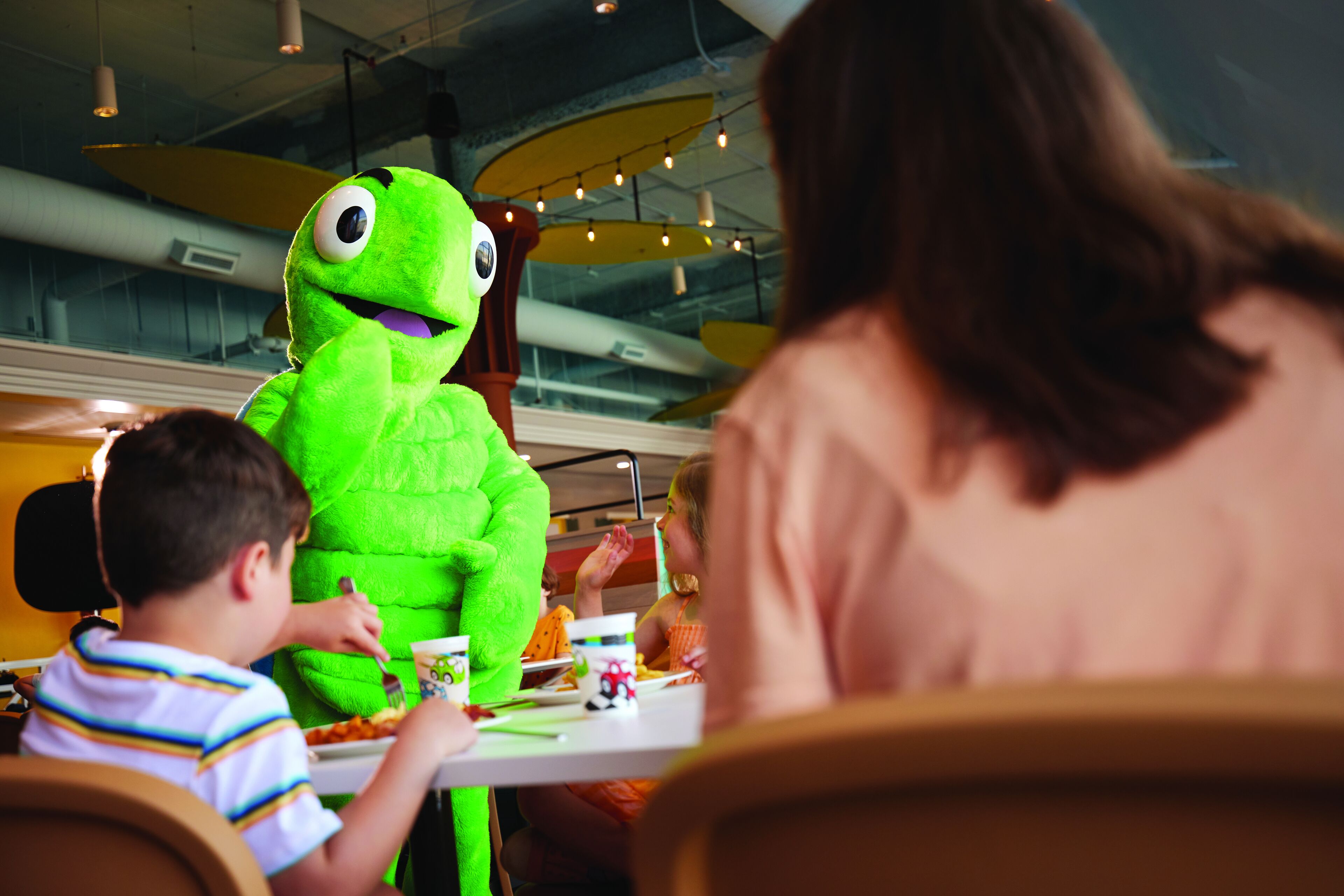 A restaurant with a green mascot, tables, chairs, and a ceiling with lights.