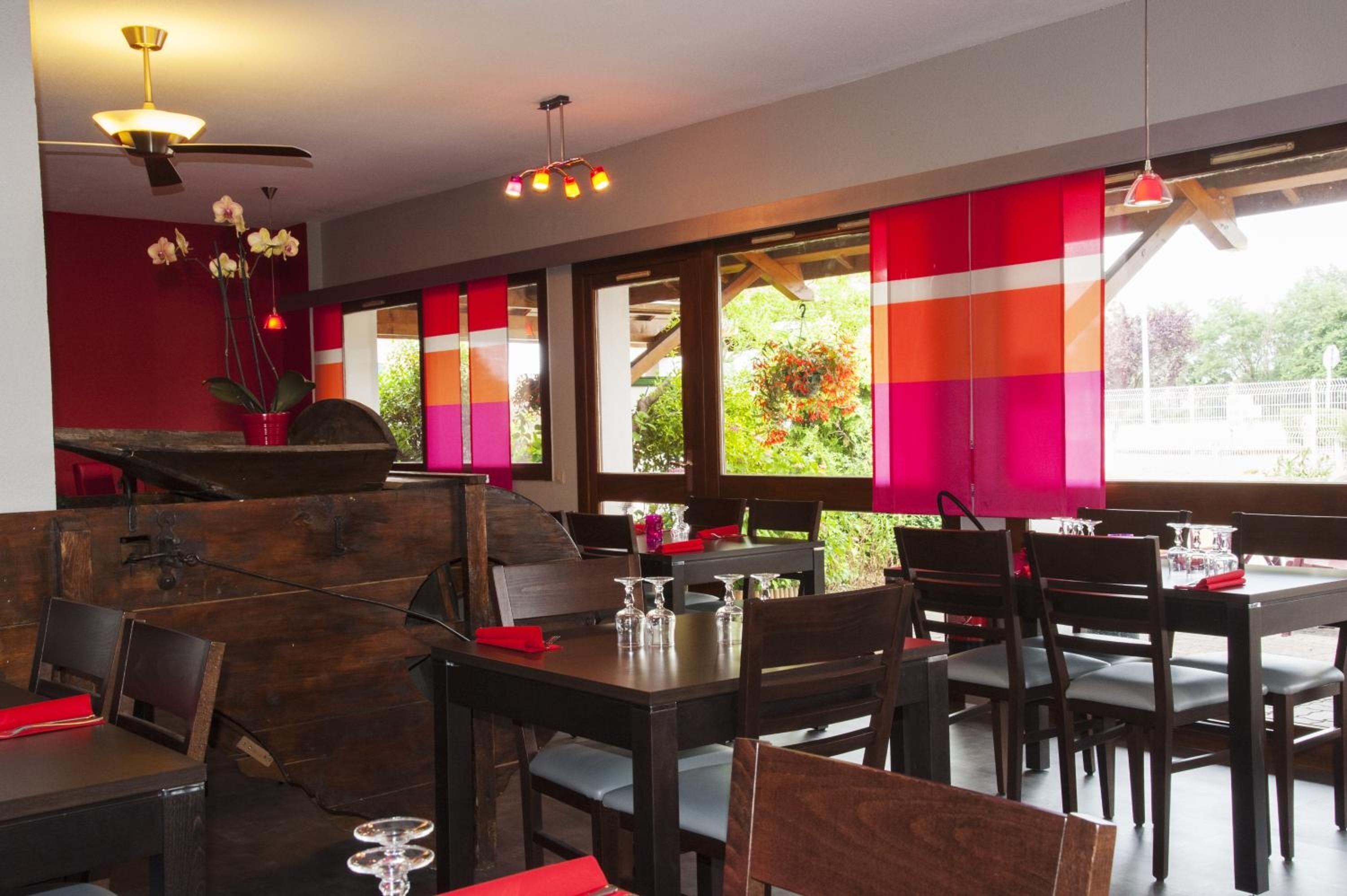 Un restaurante con mesas y sillas de madera, paredes rojas y ventanas amplias con cortinas coloridas.