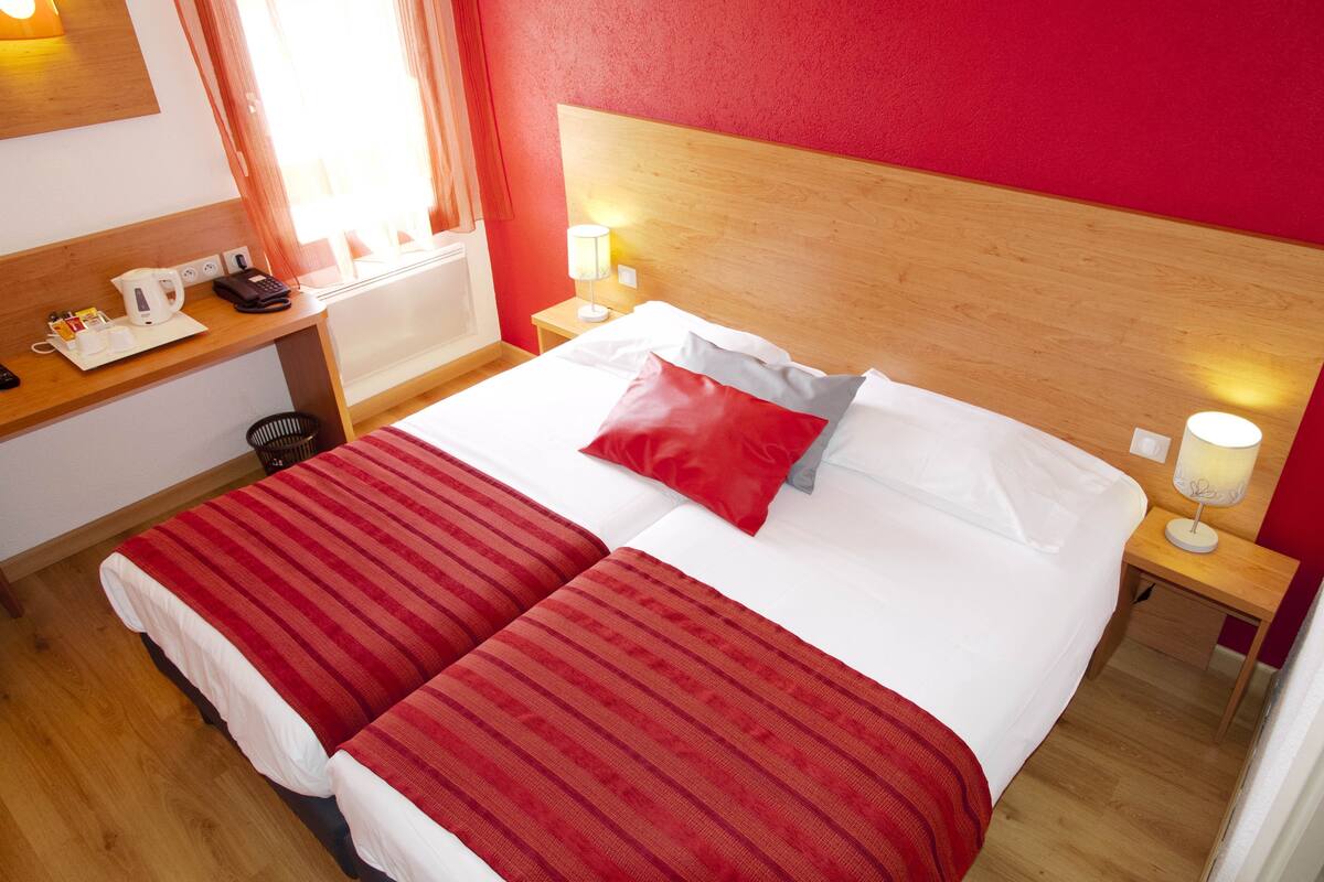A hotel room with a double bed, a wooden headboard, bedside lamps, a nightstand, a desk with a telephone, a kettle, and a basket.