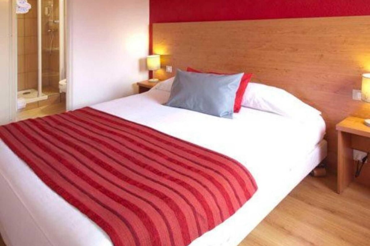 A hotel room with a bed, a wooden headboard, a red-striped throw blanket, and a nightstand with a lamp.