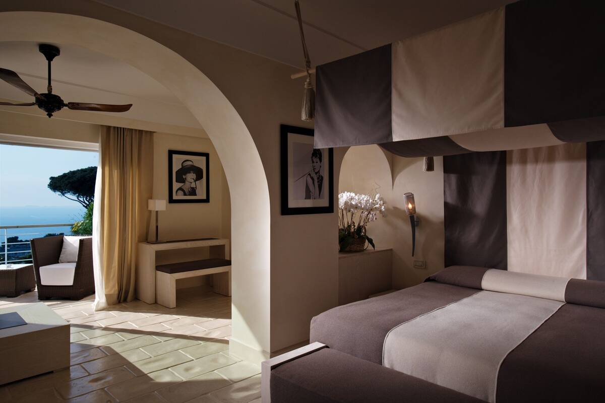 A modern hotel room with a large bed, a seating area with a sofa and chairs, a ceiling fan, and a view of the sea.