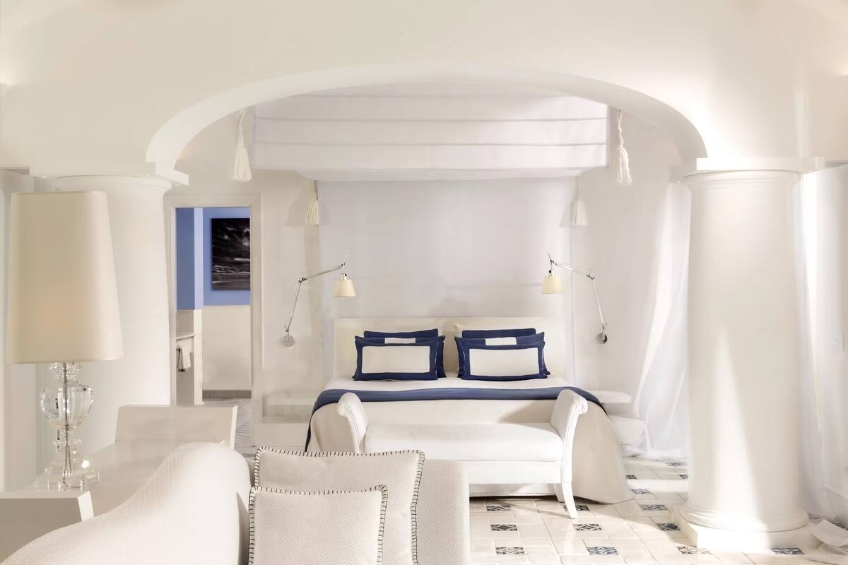A bedroom with a white bed, two bedside lamps, a white chair, and a view through an arched doorway.
