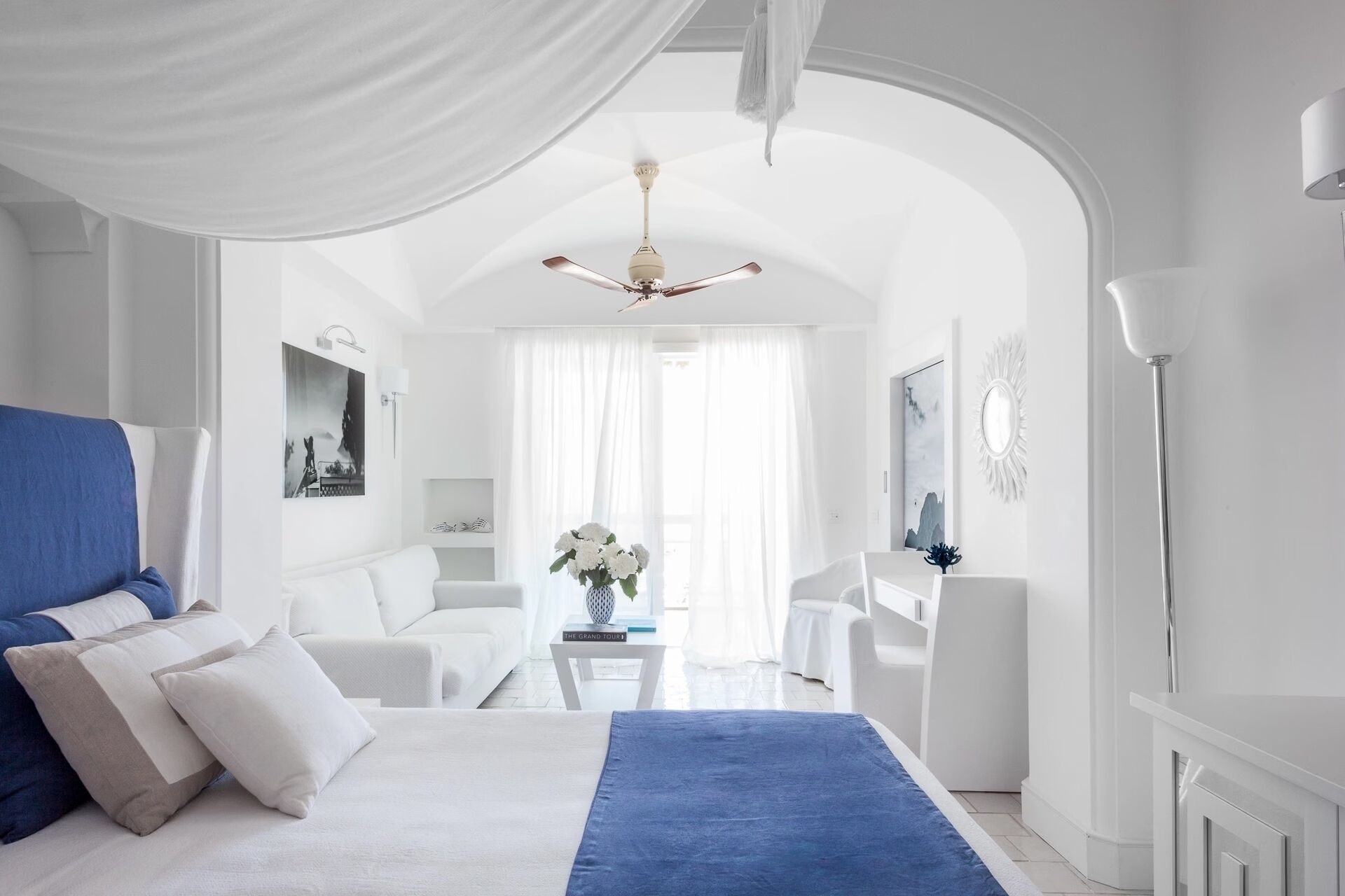 A modern hotel room with a large bed, a sitting area with a table, and a view through an arched doorway.