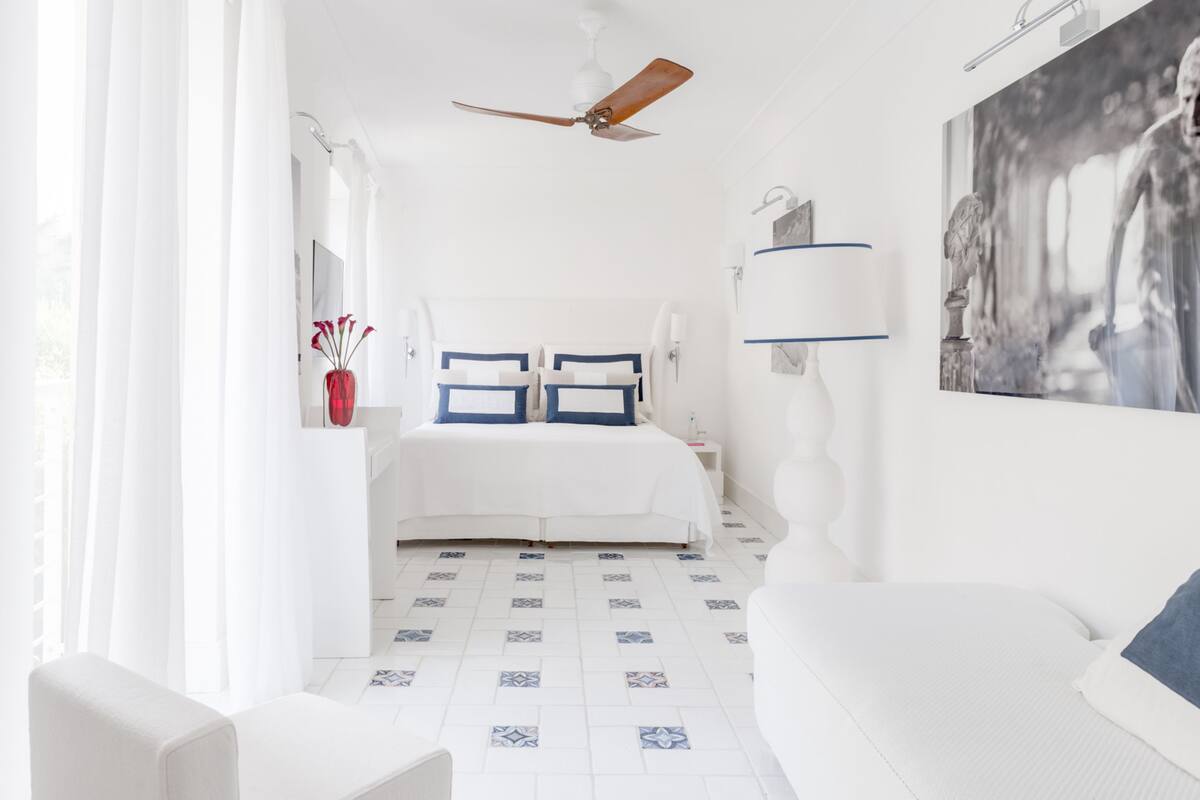 A white bedroom with a bed, a ceiling fan, a bedside table, a lamp, and a large painting on the wall.