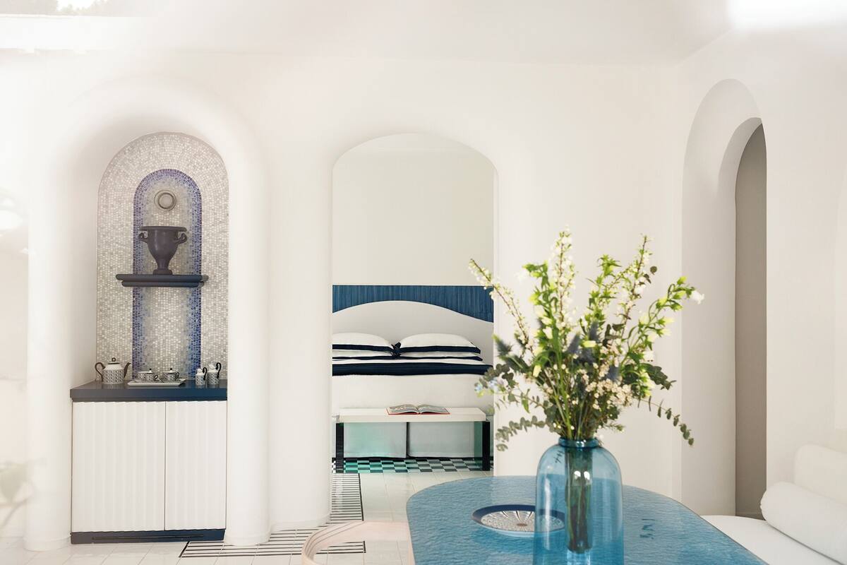 A modern hotel room with a checkered floor, a bed with striped bedding, a blue vase with flowers, and a mosaic wall.