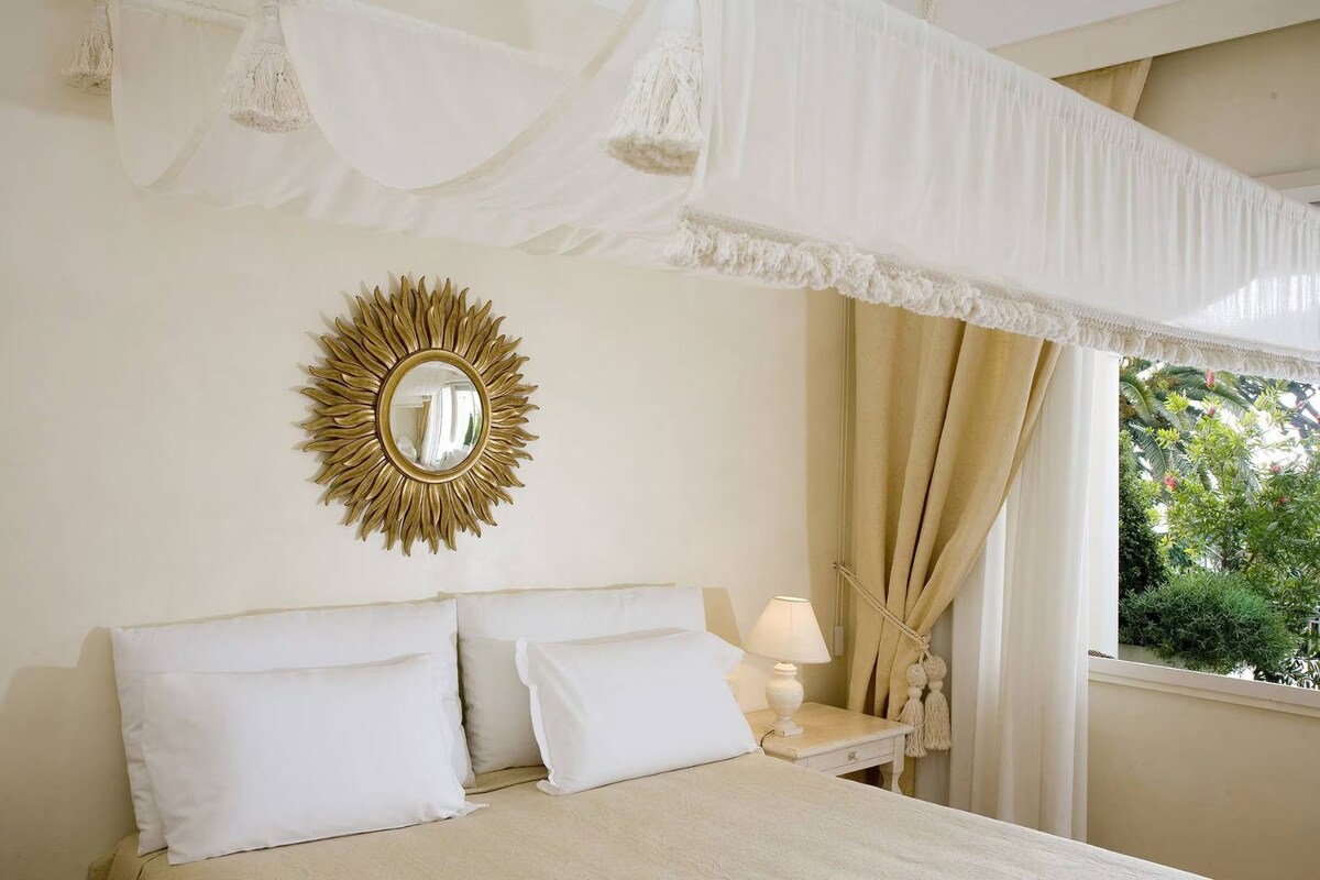 A bedroom with a bed, a nightstand, a lamp, a window with curtains, and a sunburst mirror on the wall.