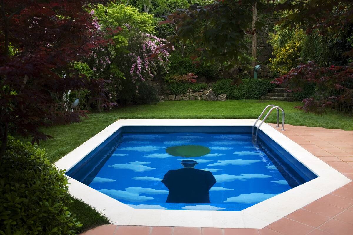 A rectangular swimming pool with a white border, surrounded by lush greenery and a stone wall.