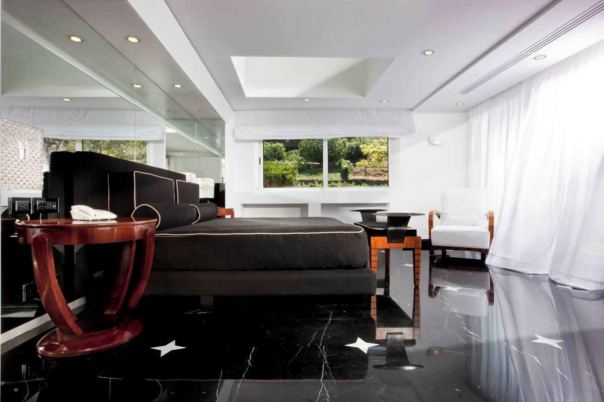 A modern hotel room with a large bed, a desk, a chair, and a view of greenery outside.