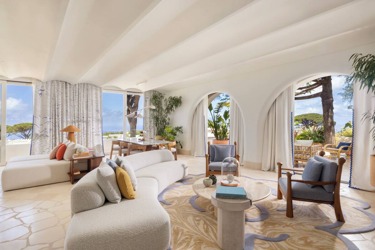 A spacious living area with a large sofa, armchairs, a coffee table, and a dining table. There are potted plants and a view of the outdoors.