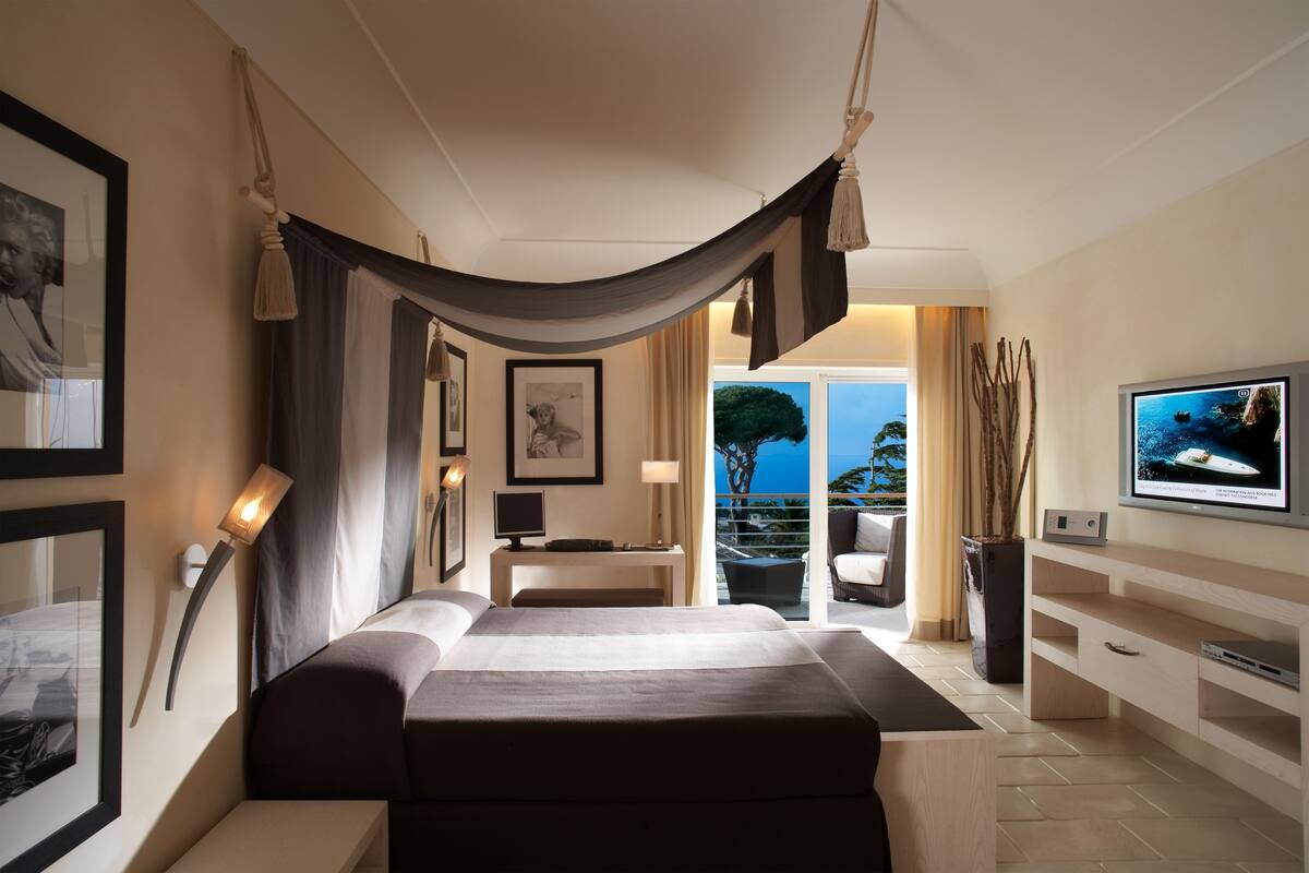 A modern hotel room with a large bed, a TV, a desk, and a balcony with a view of the sea.