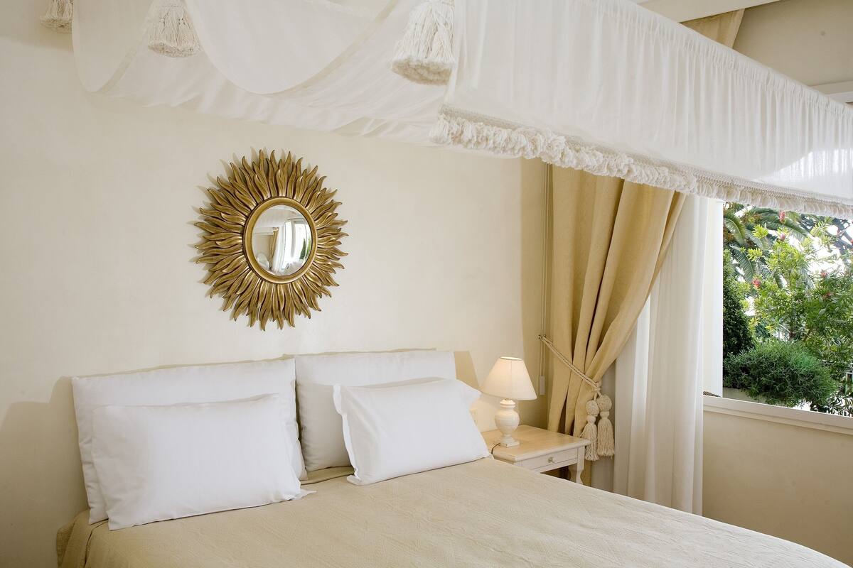 A bedroom with a canopy bed, a sunburst mirror, and a view of greenery outside.