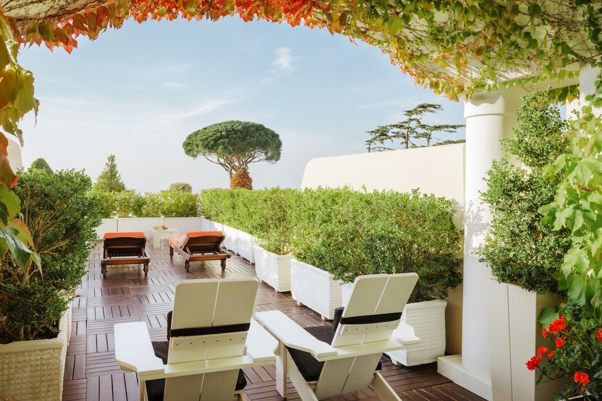 An outdoor seating area with white chairs and a wooden table, surrounded by greenery and a white wall.