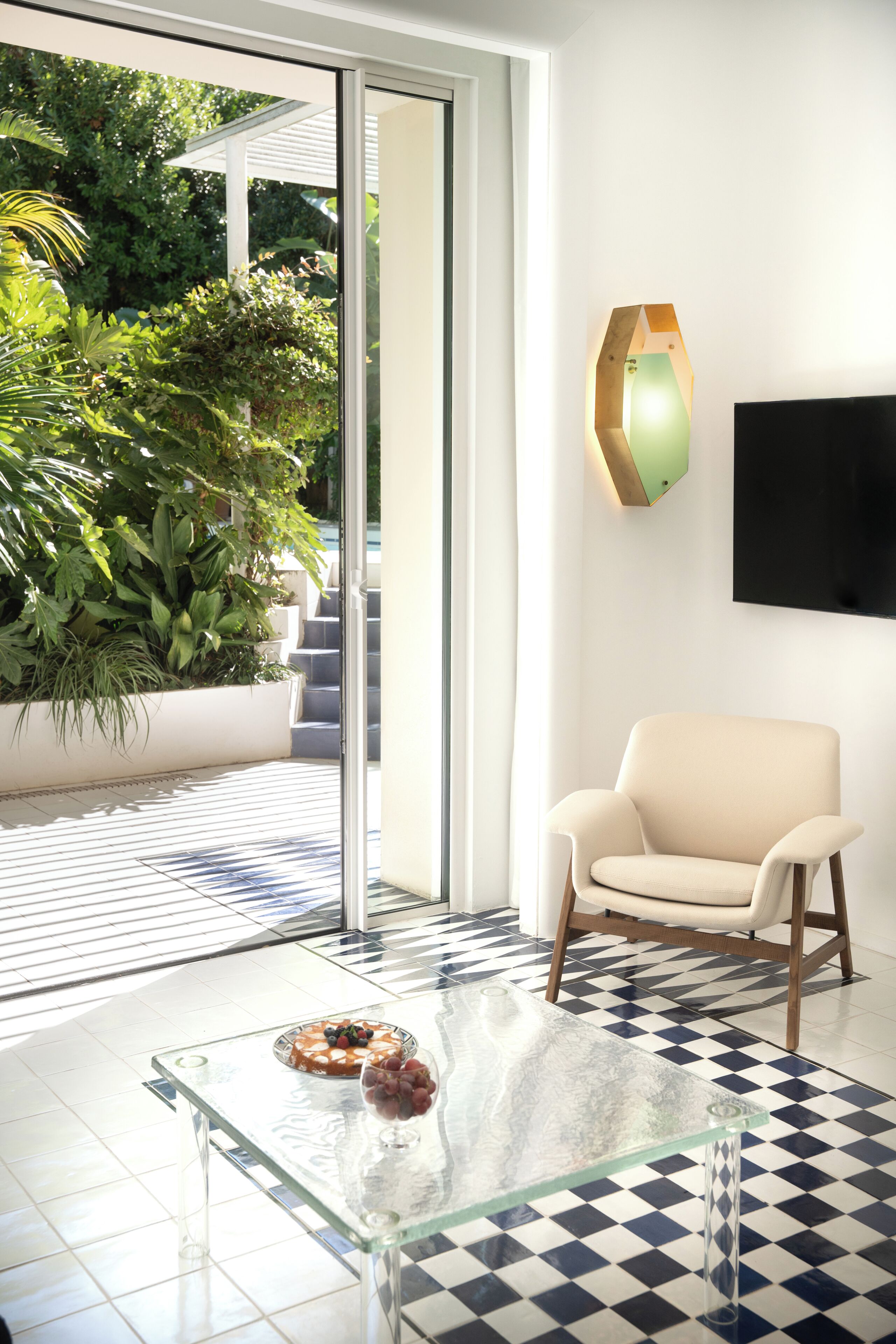 A modern room with a glass coffee table, a beige armchair, a mounted TV, and a view of a garden through a sliding glass door.
