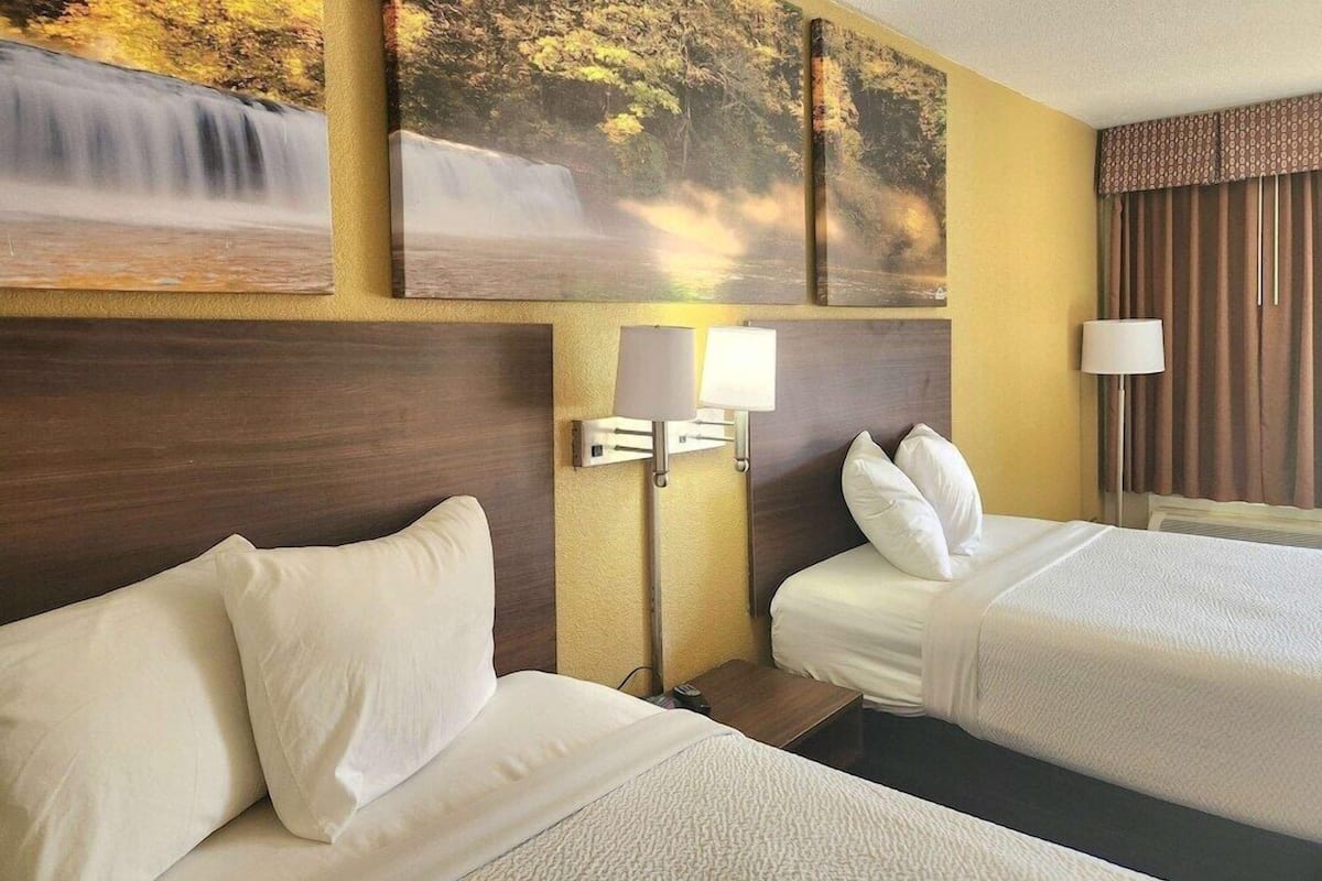 Habitación de hotel con dos camas, cabecero, mesita de noche, lámpara y un cuadro de una cascada.