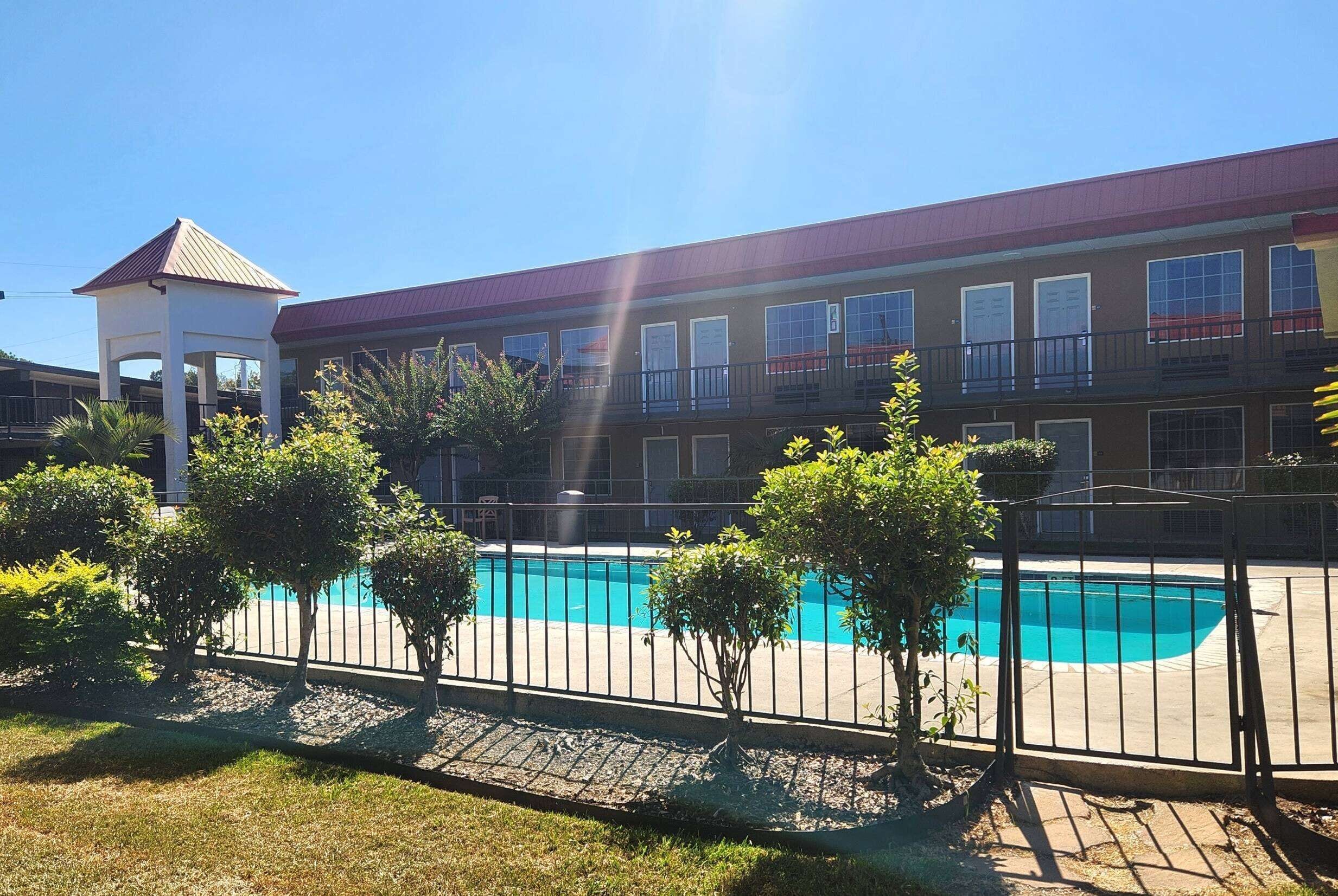 A hotel with a swimming pool, a covered gazebo, and a fenced area with trees.