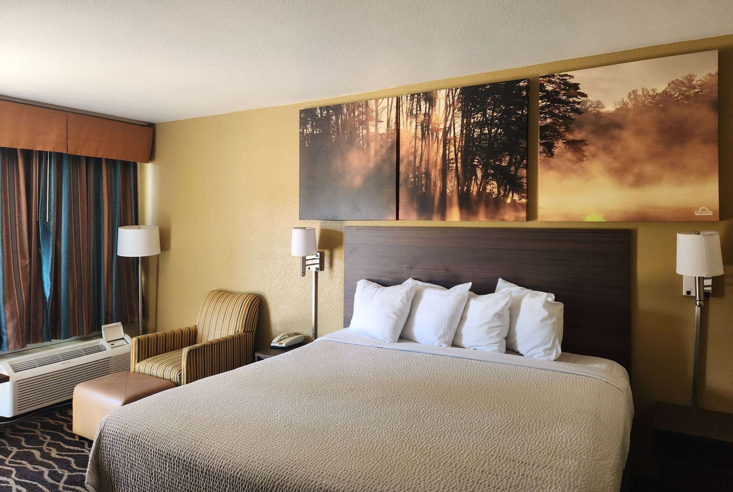 Quarto de hotel com cama grande, uma cadeira, escrivaninha e janela com cortinas.