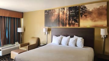 Quarto de hotel com cama grande, uma cadeira, escrivaninha e janela com cortinas.