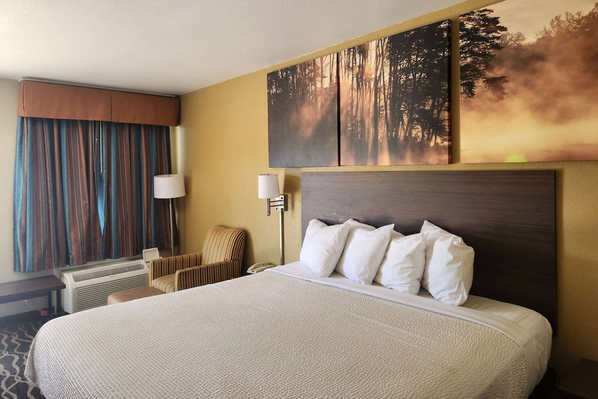 Habitación de hotel con una cama grande, cabecera, una obra de arte colgada en la pared, ventana con cortinas, una silla y un escritorio con lámpara.